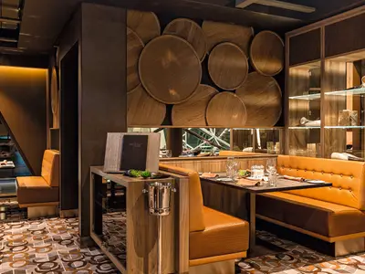 Ein elegantes Restaurant an Bord der Mein Schiff 5 mit dunklem Holz und runden Wanddekorationen. Orangefarbene Lederbänke und Tische sind einladend gedeckt. Links führt eine schräge Wand mit indirekter Beleuchtung in einen weiteren Bereich. Rechts sind beleuchtete Regale mit Dekorationsartikeln zu sehen. Der Boden ist mit einem gemusterten Teppich ausgelegt.