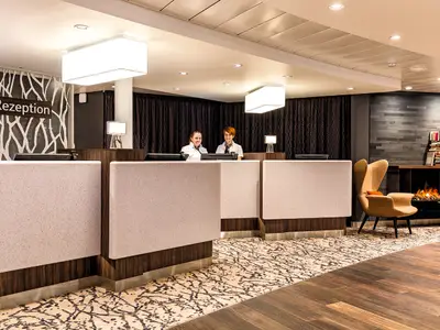 Zwei lächelnde Rezeptionistinnen hinter modernen Empfangstresen an Bord der Mein Schiff 5. Links eine Wand mit weißem Baumrindenmuster, rechts ein Lounge-Bereich mit Kamin, Sessel und Büchern. Der Boden ist mit einem gemusterten Teppich ausgelegt, der in einen Holzfußboden übergeht.