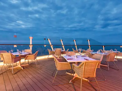 Ein Außendeckrestaurant der Mein Schiff 5 bei Sonnenuntergang. Mehrere Tische mit Rattanstühlen stehen auf einem Holzdeck. Die Tische sind mit weißer Tischwäsche, Gläsern und Besteck gedeckt. Eine Glasbrüstung bietet einen ungehinderten Blick auf das dunkle Meer und den bewölkten blauen Himmel.