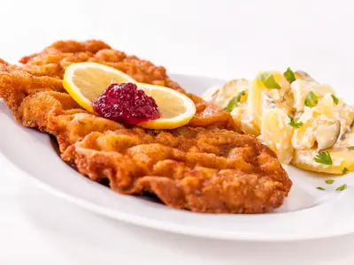 Ein appetitliches, goldbraun paniertes Wiener Schnitzel auf einem weißen Teller, garniert mit einer Zitronenscheibe und Preiselbeeren. Daneben befindet sich eine Portion cremiger Kartoffelsalat mit Gurkenstücken und frischer Petersilie. Dieses Gericht könnte auf Mein Schiff 5 serviert werden.