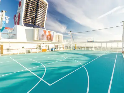 Ein Sportfeld an Bord der Mein Schiff 5, mit einem türkisfarbenen Boden und weißen Linien für Basketball. Im Hintergrund sind Teile des Schiffes und des Meeres unter einem blauen Himmel zu sehen. Ein großer Bildschirm zeigt Werbung.