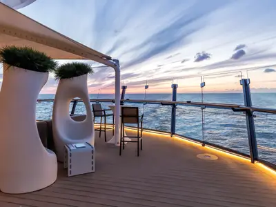 Eine Outdoor-Lounge auf der Mein Schiff 5 bei Sonnenuntergang. Der Holzdeckbereich ist mit modernen Möbeln, wie hohen Pflanzengefäßen mit grünem Gras, Sitzgelegenheiten und Bartischen, ausgestattet. Eine umlaufende LED-Beleuchtung schafft eine warme Atmosphäre. Im Hintergrund ist das weite Meer unter einem farbigen Himmel zu sehen, während die Sonne untergeht. Eine Glasreling sichert den Bereich und bietet freie Sicht.