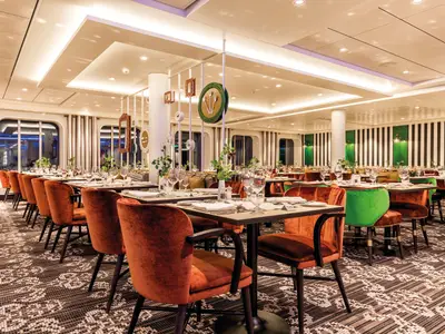 Ein elegantes, hell erleuchtetes Restaurant an Bord der Mein Schiff 5 mit vielen Tischen, gedeckt mit Geschirr und Gläsern. Die Stühle sind in leuchtendem Orange und Grün gehalten. Die Wände sind teilweise mit vertikalen Streifen in Schwarz und Weiß verziert, durchbrochen von ovalen Fenstern, die den Blick auf das Meer freigeben.