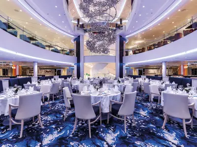 Blick in das geräumige und elegant eingerichtete Restaurant 'Atlantic' an Bord der Mein Schiff 5. Zahlreiche Tische mit weiß gedeckten Tischtüchern und Stühlen sind auf einem blauen Teppich verteilt. Im Hintergrund erstreckt sich ein offenes Atrium mit einer spiralförmigen Treppe und einer großen, schillernden Lichtinstallation in der Mitte.