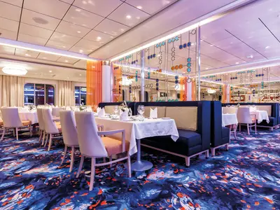 Innenansicht eines stilvollen Restaurants an Bord der Mein Schiff 5. Zahlreiche Tische mit weißen Tischdecken, modernen Stühlen und blauen Sitzbänken sind auf einem leuchtend blauen Teppich mit orangen Akzenten angeordnet. Große Fenster auf der linken Seite bieten Ausblick nach draußen. Im Hintergrund befindet sich eine dekorative Spiegelwand mit farbigen Kreisen.