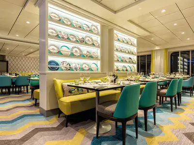 Ein helles Restaurant an Bord der Mein Schiff 5. Gelbe Sitzbänke und türkise Stühle stehen an weiß gedeckten Tischen. An der Wand sind beleuchtete Regale mit bunten Dekorativtellern. Der Teppich hat ein Wellenmuster in Grau, Gelb und Türkis.