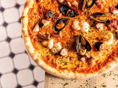 Eine appetitliche Meeresfrüchte-Pizza, reich belegt mit Garnelen, Muscheln und Tintenfisch, appetitlich angerichtet an Bord der Mein Schiff 5. Von oben aufgenommen, zeigt sie die goldbraune Kruste und den geschmolzenen Käse, bestreut mit frischen Kräutern.