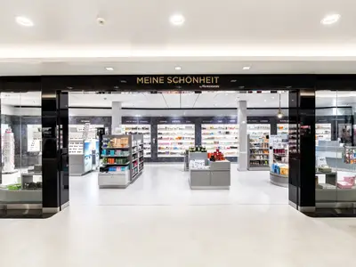 Blick in den Duty-Free-Shop "MEINE SCHÖNHEIT by HEINEMANN" auf der Mein Schiff 5, mit Kosmetik- und Parfumregalen. Links und rechts sieht man Bereiche mit Sonnenbrillen und weiteren Produkten.