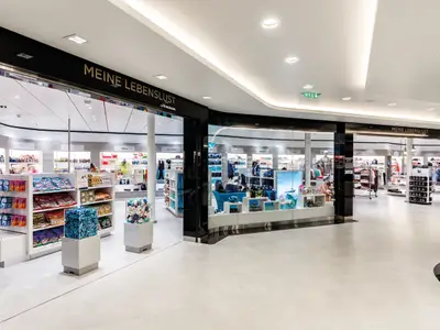 Blick in eine helle, modern gestaltete Einkaufspassage an Bord der Mein Schiff 5. Mehrere Shops mit großen Glasfronten bieten Süßigkeiten, Kleidung und Accessoires an. Der Boden ist hell gefliest.