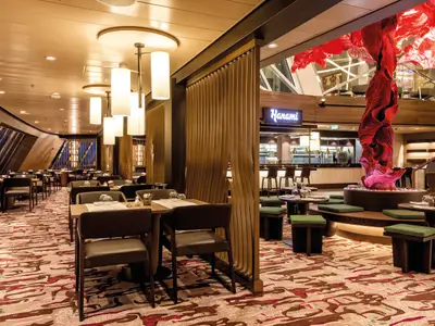Innenansicht des Restaurants Hanami auf der Mein Schiff 5. Links sind zahlreiche Tische mit Stühlen zu sehen, rechts ein Bar-Bereich und ein großes rotes Kunstwerk, das von der Decke hängt. Der Boden hat ein gemustertes Teppichdesign. Eine warme Beleuchtung schafft eine gemütliche Atmosphäre.