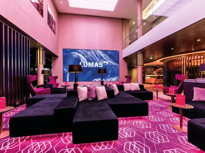 Innenansicht der Lumas Bar an Bord der Mein Schiff 5. Ein großer Lounge-Bereich mit dunklen Sofas und pinken Kissen ist zu sehen. Eine große Videowand zeigt blaue Wassermotive und den Schriftzug LUMAS. Der Boden ist mit einem pinken Teppich mit geometrischen Mustern ausgelegt. Im Hintergrund ist eine Bar zu sehen.
