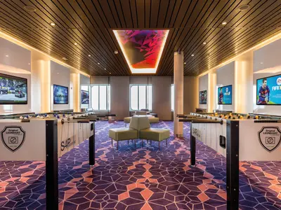 Ein geräumiger Entertainment-Raum an Bord der Mein Schiff 5 mit mehreren Kickertischen, großen Bildschirmen, auf denen Renn- und Fußballspiele laufen, sowie bequemen Sitzgelegenheiten in der Mitte und einem auffälligen Deckenlicht in der Form eines abstrakten Kunstwerks.