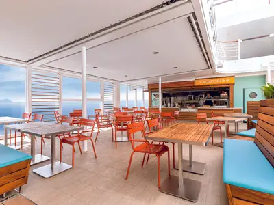 Blick in das offene, helle Selbstbedienungs-Restaurant "Bosporus" an Bord der Mein Schiff 5. Zahlreiche orangefarbene Stühle und Holztische sind zu sehen. Links große Fenster mit Blick auf das blaue Meer und eine bergige Küstenlinie in der Ferne. Rechts eine Holzeinrichtung mit blauen Sitzkissen und grünen Pflanzen. Im Hintergrund ist die Servicetheke des Restaurants zu sehen.