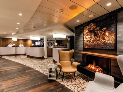 Die Rezeption der Mein Schiff 5 mit einem hell erleuchteten Kunstwerk über einem Gaskamin. Zwei Empfangsmitarbeiter und zwei Gäste sind zu sehen. Der Boden wechselt von dunklem Holz zu einem hell gemusterten Teppich. Eine beigefarbene Ohrensessel und eine kleine Ablage stehen im Vordergrund. An der Wand hängen Uhren.