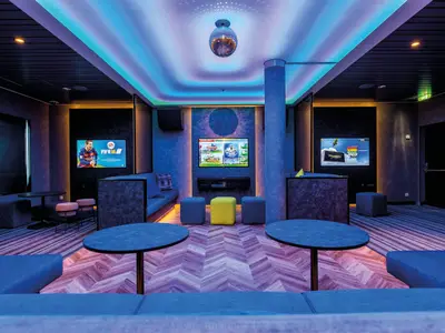 Innenansicht eines farbenfroh beleuchteten Spielraums an Bord der Mein Schiff 5. In der Mitte des Raumes befinden sich mehrere Fernseher, auf denen Videospiele laufen. Blaue Sofas und Tische sind im Vordergrund und an den Seiten angeordnet.