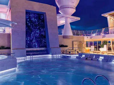 Blick auf das obere Deck der Mein Schiff 5 bei Nacht, mit einem großen, blau beleuchteten Pool im Vordergrund, einem Jacuzzi links und Liegestühlen. Im Hintergrund sind Teile der Schiffsarchitektur und eine blau leuchtende Wand zu sehen.
