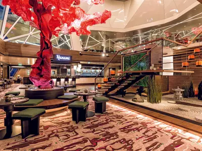 Ein luxuriöses japanisches Restaurant namens Hanami an Bord der Mein Schiff 5. Links ein roter Baumstamm mit Blättern, in der Mitte Tische und Stühle, rechts ein Zen-Garten mit Steinen und Pflanzen und einer Treppe.