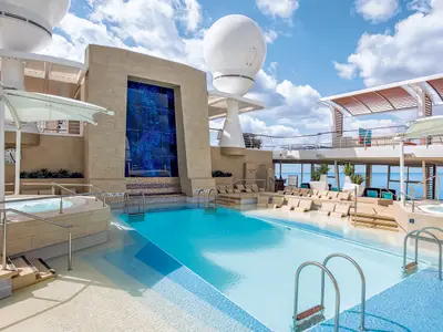 Blick auf den weitläufigen Außenpoolbereich an Deck der Mein Schiff 5. Das rechteckige Poolbecken mit Liegestühlen im flachen Wasser ist umgeben von weiteren Liegen und einem Whirlpool. Im Hintergrund ist das offene Meer unter blauem Himmel mit weißen Wolken zu sehen.