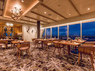 Blick in das elegant eingerichtete Restaurant Schmankerl an Bord der Mein Schiff 5. Mehrere Holztische mit gedeckten Plätzen und bequemen Stühlen stehen auf einem gemusterten Teppich. An der Decke hängt ein großer Hirschgeweih-Kronleuchter. Große Fenster bieten einen Blick auf das blaue Meer.