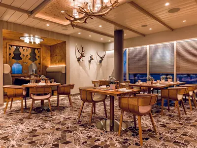 Ein elegantes Restaurant auf der Mein Schiff 5 mit Holztischen und passenden Stühlen, Hirschgeweihen an der Wand und großen Fenstern mit Jalousien, die einen Blick auf das Meer freigeben.