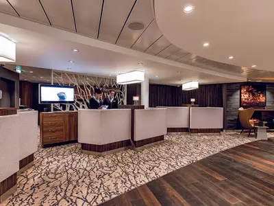 Blick auf die Rezeption der Mein Schiff 5. Zwei männliche Rezeptionisten stehen hinter den hellen Schaltern mit Holzverkleidung. Hinter ihnen befindet sich eine Wand mit einem stilisierten Baumdesign und der Aufschrift "Rezeption". Im Hintergrund ist ein großer Fernseher zu sehen, der ein Schiff zeigt. Rechts laden bequeme Sessel zum Entspannen ein.