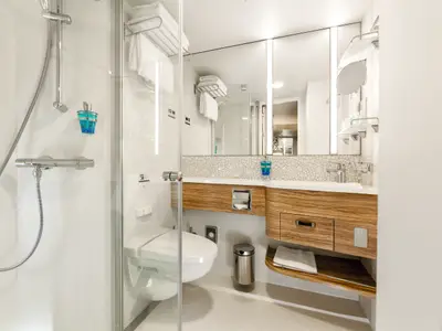 Helles, modernes Badezimmer an Bord der Mein Schiff 5 mit Duschkabine, WC mit Bidet, Waschtisch mit Spiegel und Holzschrank. Ordentlich gefaltete Handtücher liegen bereit.