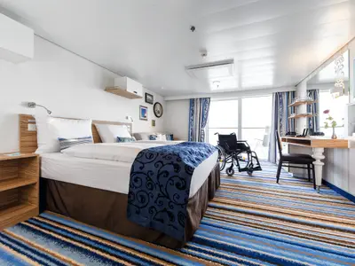 Eine helle, barrierefreie Balkonkabine an Bord der Mein Schiff 5. Im Vordergrund ein gemütliches Bett mit blauer Tagesdecke, dahinter ein Schreibtisch und ein Rollstuhl vor dem Balkonfenster.