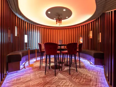 Blick in die elegante Galerie Bar an Bord der Mein Schiff 5. Rote Samtstühle und eine geschwungene Sitzbank umgeben einen Tisch. Indirekte Beleuchtung in Orange und Lila schafft eine moderne, einladende Atmosphäre in diesem Innenbereich.
