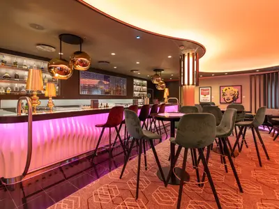 Die moderne Galerie Bar an Bord der Mein Schiff 5. Der Tresen leuchtet pink, davor stehen samtige Barhocker. Im Hintergrund sind Spirituosenregale und Kunstwerke zu sehen.