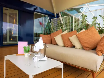 Ein Champagner-Treff an Bord der Mein Schiff 5. Ein weisser Tisch mit Champagnerflasche im Kuehler, Glaesern und einer Karte steht vor einem bequemen Sofa mit orange-beigen Kissen auf dem Aussendeck.