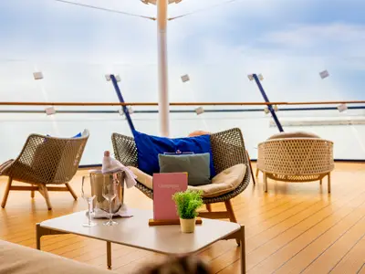 Eine Terrasse an Bord der Mein Schiff 5 zeigt bequeme Loungesessel, einen Tisch mit Champagnerkühler, zwei Gläsern und einer Speisekarte. Im Hintergrund ist das Meer unter hellem Himmel zu sehen.