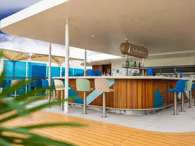 Die moderne Außenalster Bar an Bord der Mein Schiff 5. Sie zeigt eine Holztheke mit blauen Mustern, bunte Barhocker und dahinter den Poolbereich unter blauem Himmel.