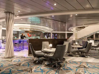 Ein Barkeeper bereitet Drinks in der TUI Bar an Bord der Mein Schiff 5 zu. Moderne Lounge-Sessel und Tische auf einem Teppich. Elegante Inneneinrichtung mit Treppe im Hintergrund.