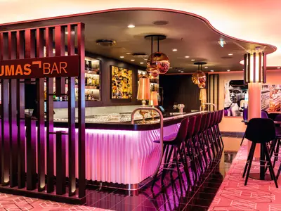 Innenansicht der Lumas Bar an Bord der Mein Schiff 5. Die Barfront ist mit vertikalen Leisten und lila Beleuchtung versehen. Daneben stehen dunkle Barhocker. Im Hintergrund sind Regale mit Flaschen und Kunstwerke zu sehen, darunter ein Porträt von Muhammad Ali. Die Decke ist geschwungen und mit Einbaustrahlern versehen.