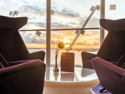 Zwei moderne, dunkle Sessel rahmen einen runden Glastisch ein. Darauf stehen eine Vase mit einer stilisierten Blume und eine braune "Lounge"-Menükarte. Im Hintergrund ist ein großer Fensterbereich zu sehen, der einen goldenen Sonnenuntergang über dem Meer offenbart. Es handelt sich um eine Szene an Bord der Mein Schiff 5.