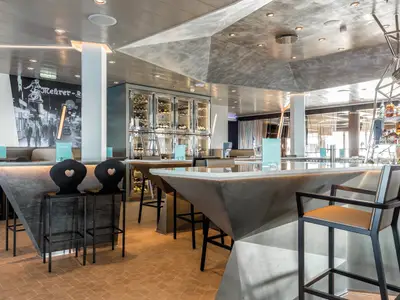 Ein weitwinkliges Innenansicht einer modernen Bar auf der Mein Schiff 5, die einen Barbereich mit Sitzgelegenheiten, dekorative Beleuchtung, eine Weinwand und eine große Wandillustration zeigt.