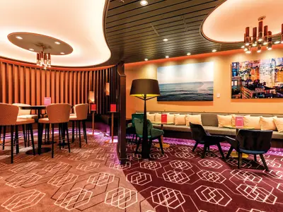 Ein stilvoller Barbereich auf der Mein Schiff 5. Links Bartische mit Hockern, rechts eine gemütliche Lounge mit Sofas und Sesseln, dekoriert mit Kunstwerken und indirekter Beleuchtung. Der Teppich hat ein geometrisches Muster.