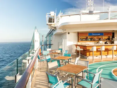Ein sonnenbeschienener Außenbereich an Bord der Mein Schiff 5 mit einer voll ausgestatteten Bar im Hintergrund und mehreren Tischen und Stühlen im Vordergrund. Links ist das offene Meer unter blauem Himmel sichtbar.