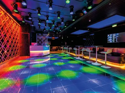Ein breiter Blick in die futuristische LUMAS Bar der Mein Schiff 5. Der Raum ist in dunklen Tönen gehalten, mit einer beleuchteten Bar, stylischen Hockern an Tischen und einer gemütlichen Sitzbank. Die Tanzfläche leuchtet in leuchtenden Farben, die von roten und blauen Wänden reflektiert werden.