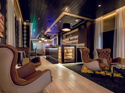 Ein luxuriöser Barbereich an Bord der Mein Schiff 5. Braune Ledersessel mit hellen Akzenten stehen auf einem schwarzen Teppich mit goldenem Muster. Im Hintergrund eine gut bestückte Bar mit leuchtenden Regalen und einer Vitrine für Zigarren.
