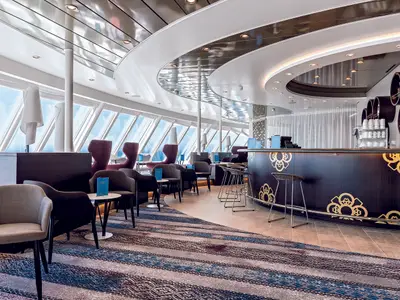 Ein breiter Blick auf eine moderne Bar-Lounge an Bord der Mein Schiff 5. Links sind große, schräge Fenster mit Blick auf das Meer zu sehen. Im Vordergrund sind bequeme Sessel und kleine Tische mit blauen Menükarten angeordnet. In der Mitte befindet sich eine geschwungene Bar in dunklem Holz mit goldenen Ornamenten. Barhocker stehen davor. Im Hintergrund ist eine helle, moderne Decke mit indirekter Beleuchtung und reflektierenden Oberflächen zu erkennen.