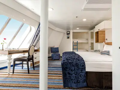 Geräumige Familienkabine Außen an Bord der Mein Schiff 5. Heller Raum mit Schreibtisch am Fenster, Doppelbett und separaten Etagenbetten. Blauer Teppich und Meerblick.