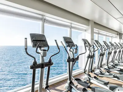 Ein Fitnessraum an Bord der Mein Schiff 5. Zahlreiche Crosstrainer stehen in einer Reihe vor großen Fenstern, die einen weiten Blick auf das offene Meer ermöglichen. Der Himmel ist hellblau und das Wasser türkisblau.
