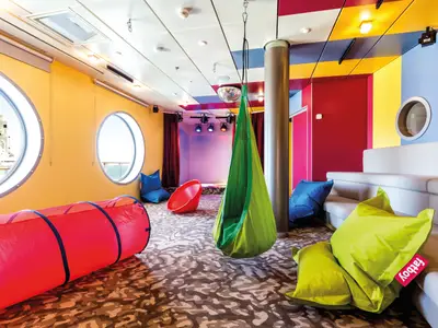 Ein farbenfroher Raum an Bord der Mein Schiff 5, gestaltet als Kinderlounge. Der Raum ist in Gelb, Rot und Blau gehalten, mit runden Bullaugen, durch die man das Meer sieht. Am Boden liegen ein roter Spieltunnel und mehrere blaue und limettengrüne Fatboy-Sitzsäcke. Eine grüne Schaukel hängt von der Decke, daneben eine rote Sitzschale. Im hinteren Bereich befindet sich eine kleine Bühne mit Scheinwerfern und einem Discokugel. An der rechten Wand steht eine hellgraue Bank.
