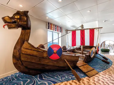 Ein großes, detailgetreues Holzmodell eines Wikingerschiffs mit Drachenkopf und Segel, das den Kinderbereich an Bord der Mein Schiff 5 schmückt. Der Boden ist mit einem Wellenmuster gestaltet und im Hintergrund sind Fenster mit Meerblick sowie ein weiterer Spielbereich zu sehen.