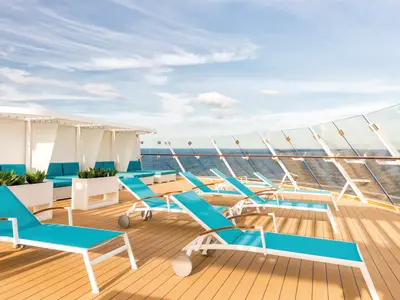 Sonnendeck der Mein Schiff 5 mit zahlreichen türkisen Liegestühlen auf hellem Holz. Im Hintergrund sind Lounges mit Polstern und Pflanzen in weißen Blumenkästen zu sehen. Die Balustrade aus Glas und die weite offene See unter einem blauen Himmel vervollständigen die Szene.