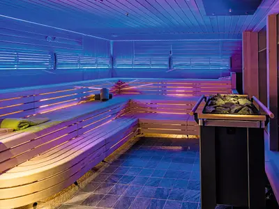Ein breiter Blick in die Sauna an Bord der Mein Schiff 5. Die Saunabänke aus hellem Holz sind mehrstufig angeordnet und werden von pinken und orangen LED-Streifen beleuchtet. Die Decke und obere Wandbereiche sind in einem kräftigen Blau getaucht. Auf einer der Bänke liegt ein grün-gelbes Handtuch. Rechts im Vordergrund steht ein moderner Saunaofen mit Lavasteinen. Der Boden ist mit dunklen Fliesen ausgelegt.