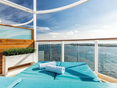 Sonnige Balkonkabine an Bord der Mein Schiff 5 mit zwei blauen Liegen und gerollten Handtüchern. Rechts ist eine Glasbrüstung mit Holzhandlauf, die den Blick auf das blaue Meer und eine grüne Küstenlandschaft freigibt. Links ist eine hölzerne Wand mit einem Pflanzenkübel.