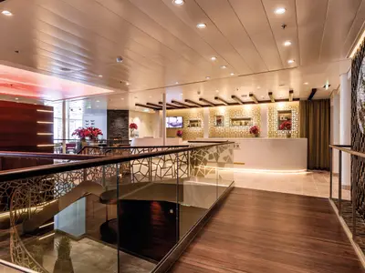 Blick in den hell erleuchteten Empfangsbereich der Mein Schiff 5. Eine weiße Rezeption mit goldenen Akzenten steht im Hintergrund. Links befindet sich ein roter Wolkenhimmel an der Decke und eine Sitzecke. Ein stilvoller Treppenaufgang mit Metallgeländer und Glasfront ist im Vordergrund zu sehen.