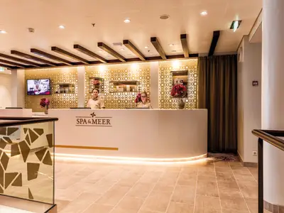 Zwei lächelnde Mitarbeiter, ein Mann und eine Frau, stehen hinter der Rezeption des "SPA & MEER" auf der Mein Schiff 5. Die Rezeption ist hell beleuchtet, dahinter goldene Wandverkleidung und Regale mit Produkten.